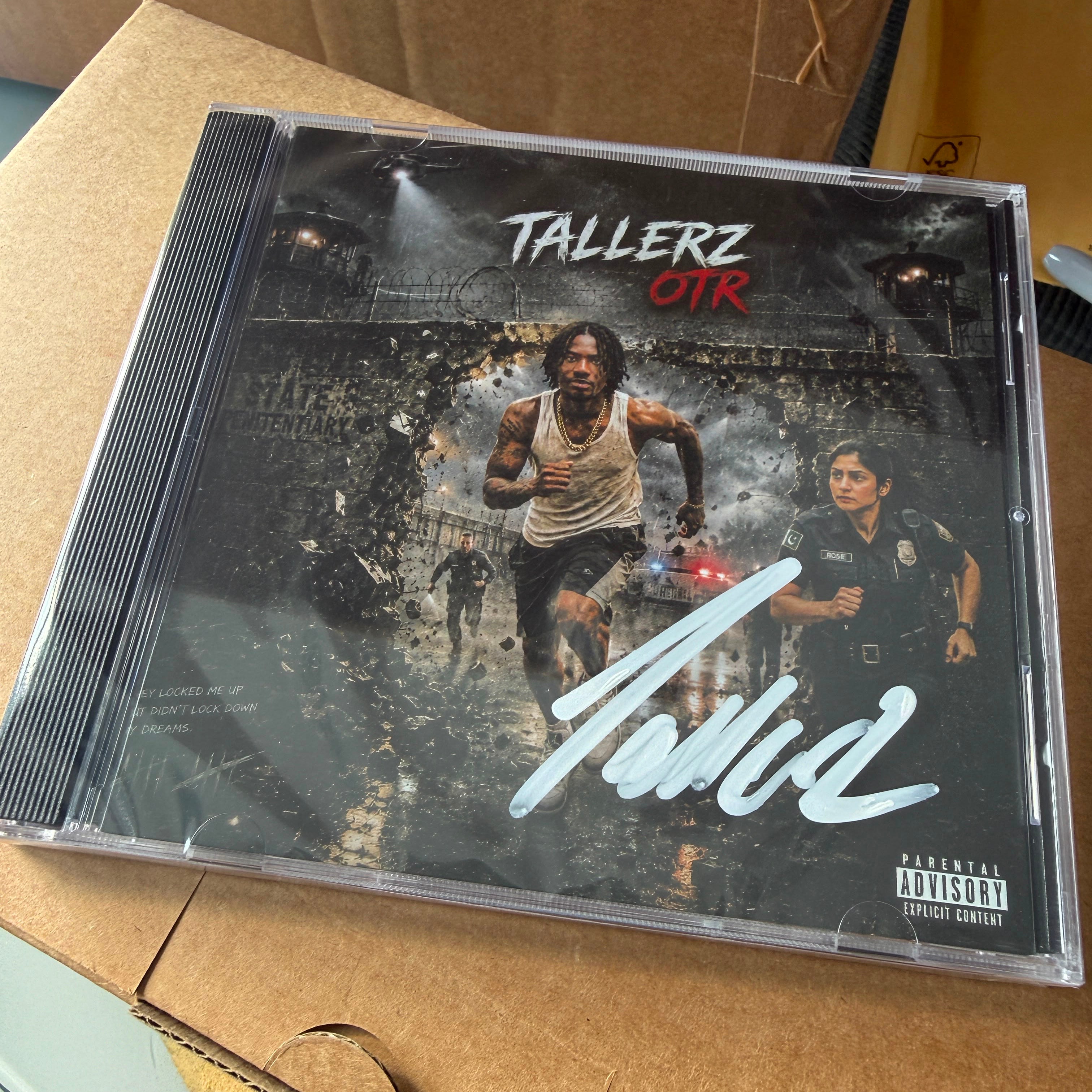 A CD case for the album 'Tallerz OTR' by the artist Tola, resting on a brown cardboard box. The album cover features a dark, gritty scene with two men, one in a tank top and one in a police uniform, with a large white signature across the front and a Parental Advisory label in the bottom right corner.