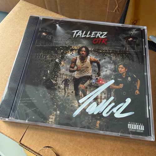 A CD case for the album 'Tallerz OTR' by the artist Tola, resting on a brown cardboard box. The album cover features a dark, gritty scene with two men, one in a tank top and one in a police uniform, with a large white signature across the front and a Parental Advisory label in the bottom right corner.